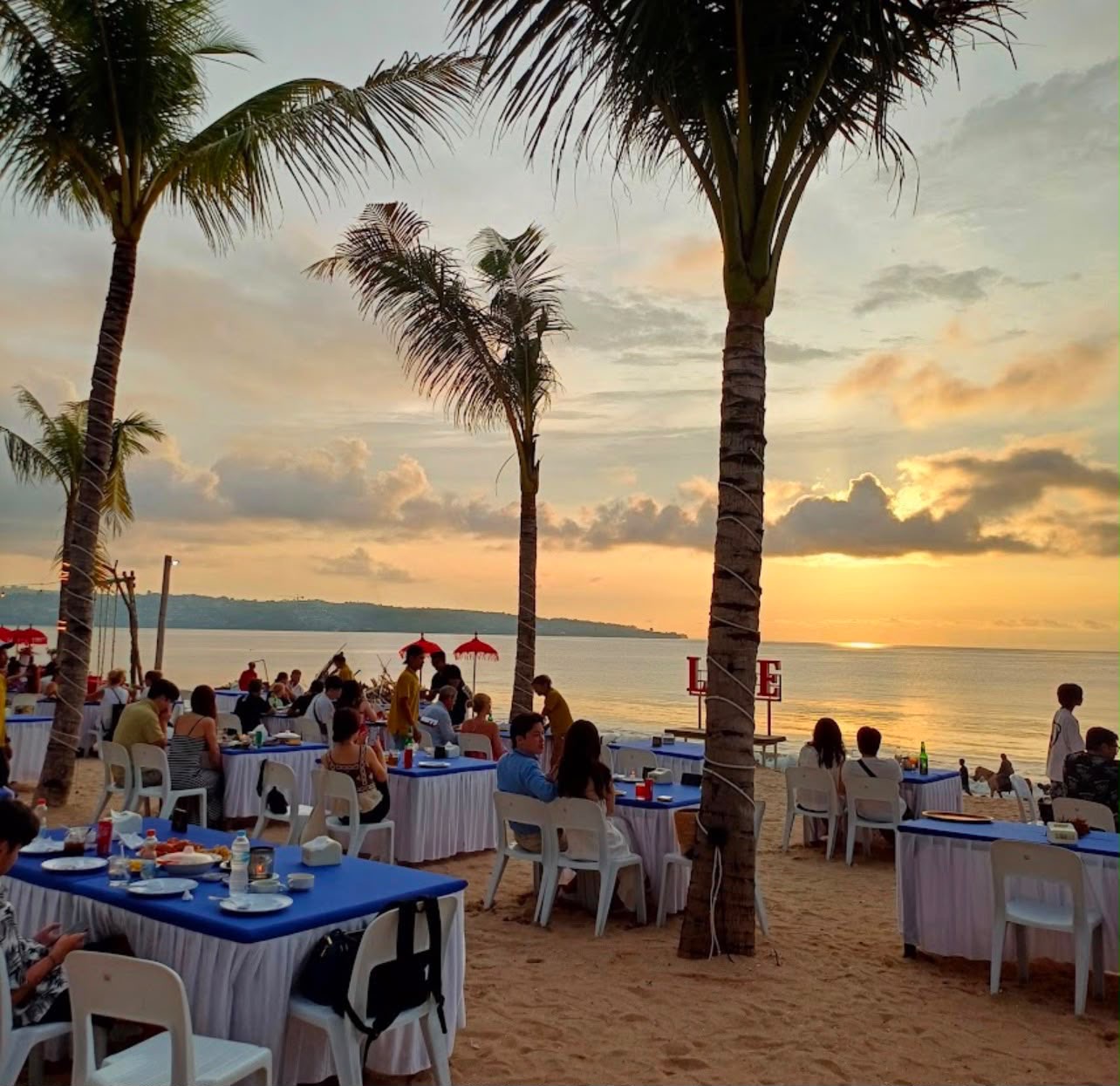Guests dining at beach tables under palm trees at sunset on Jimbaran beach